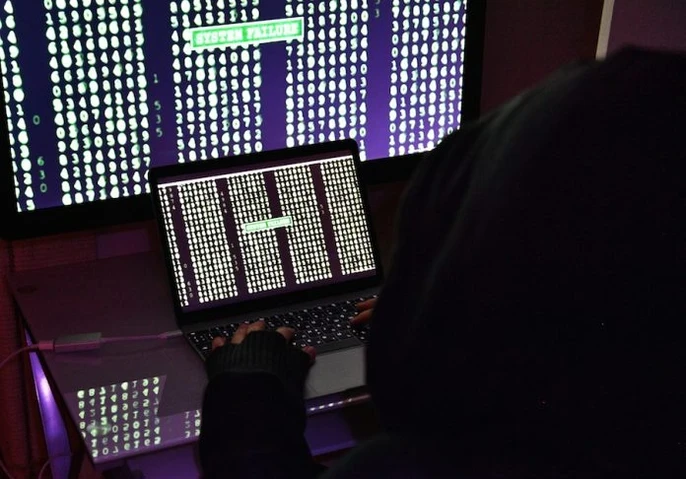 Hacker in a black hoodie sitting in front of two computer screens with lines of code.