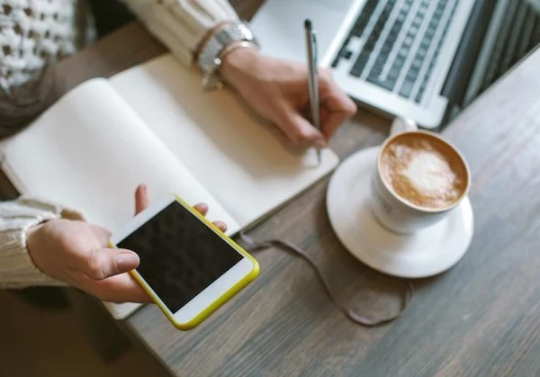 person working on a smartphone, making notes in a book with a coffee and laptop beside them