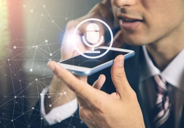 Image of someone speaking into a smartphone for voice to text with a microphone logo above the smartphone to highlight voice recognition