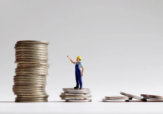 a toy worker in a hardhat standing on a small pile of coins pointing to a larger pile of coins