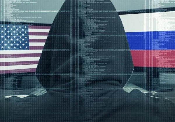 Back of a hacker in a black hoodie looking at two screens, one with the USA flag and the other with the Russia flag