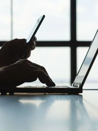 A person working on a laptop while holding a smartphone