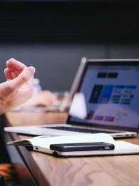 Two people in a meeting room sitting at a wooden desk with laptops and notes in front of them.