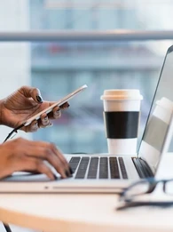 Person at a desk typing on a laptop keyboard and holding a smartphone