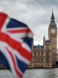 Header graphic showing the Palace of Westminster and Big Ben in London under cloudy skies, with a blurred UK flag on the left and a blurred US flag on the right. A dark translucent banner across the lower half displays the EM360Tech logo and the headline: "UK and US Authorities Increase Pressure on AI Developers After Rogue Agent Incidents".