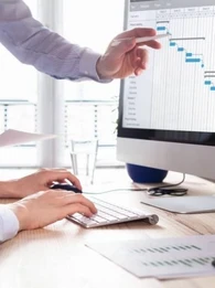 Two businesspeople standing and sitting in front of a desktop computer viewing analytics on a screen.
