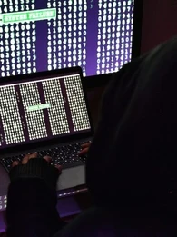 Hacker in a black hoodie sitting in front of two computer screens with lines of code.