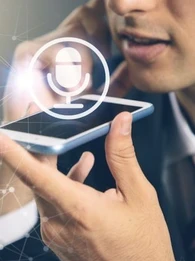 Image of someone speaking into a smartphone for voice to text with a microphone logo above the smartphone to highlight voice recognition