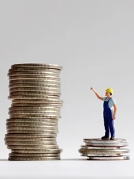 a toy worker in a hardhat standing on a small pile of coins pointing to a larger pile of coins