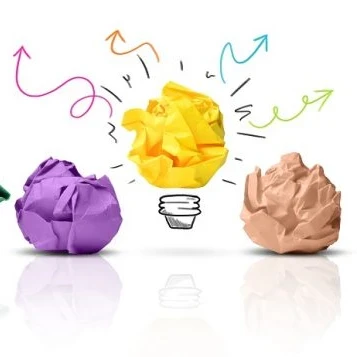 Colourful rolled up balls of paper laid out on a white table with one turning into a lightbulb.