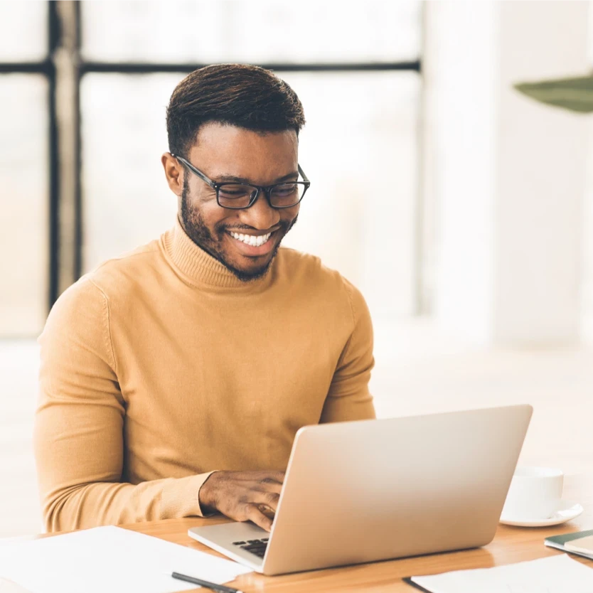 A man in a beige turtleneck sitting at a desk and working on a laptop.
