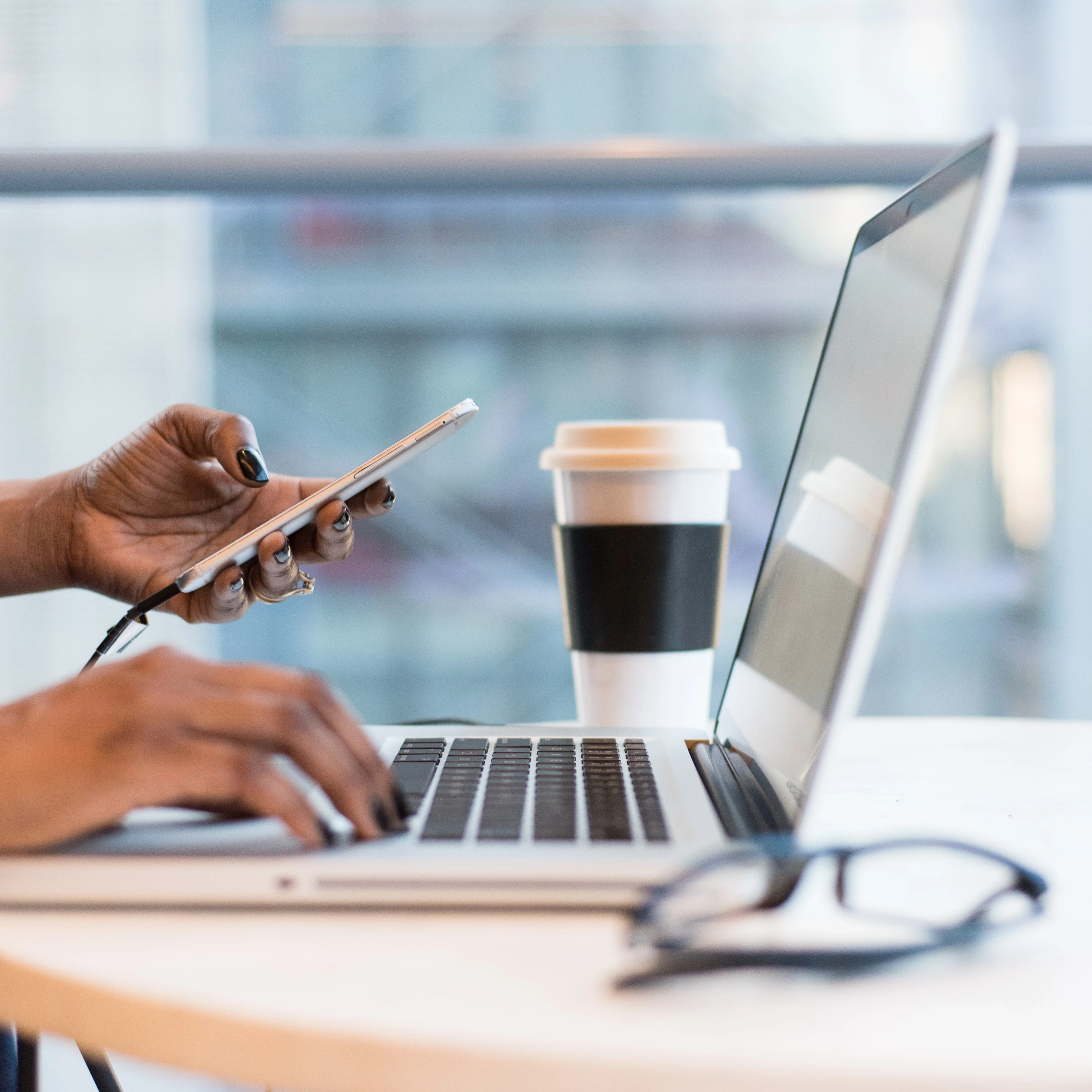 Person at a desk typing on a laptop keyboard and holding a smartphone