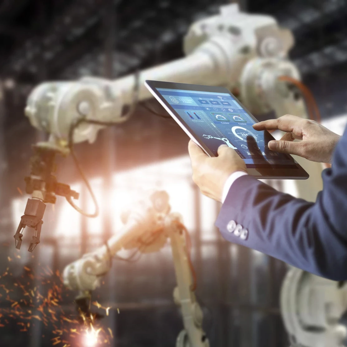 Man in a suit tapping on a tablet while a manufacturing robot works in the background.