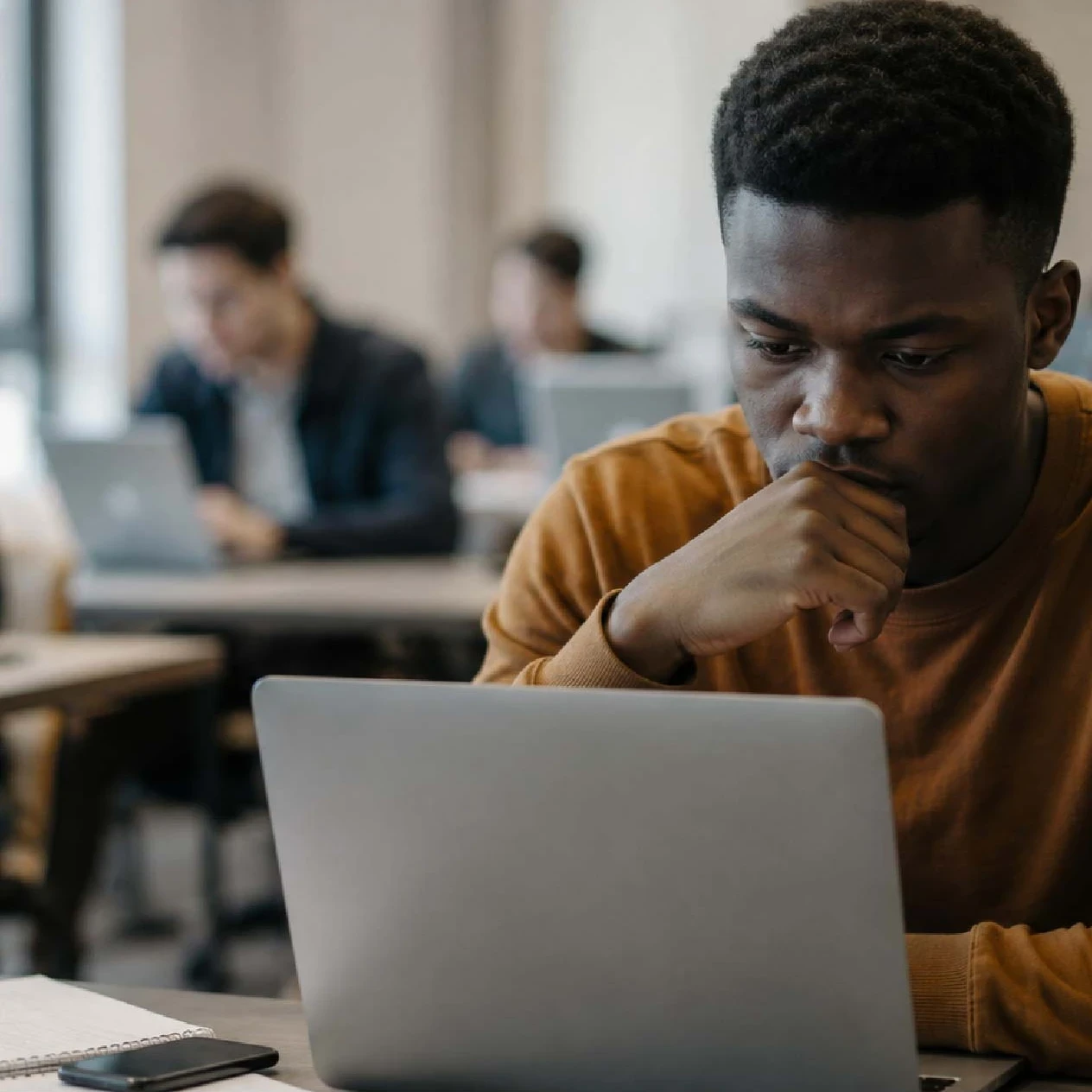 EM360Tech news graphic showing a young man looking thoughtfully at a laptop in a shared workspace, with other people working on laptops in the blurred background. Text reads: “The Youth Unemployment Crisis Is Worsening as AI Reshapes Work.” 