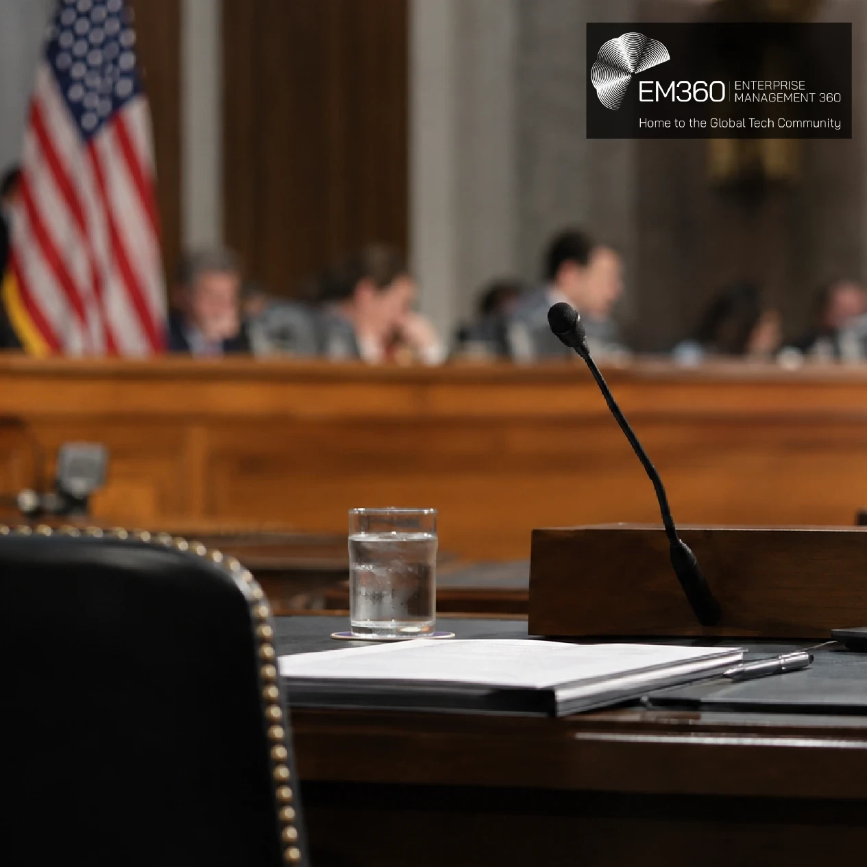 EM360Tech header graphic with the headline “OpenAI Says Voluntary AI Safety Rules Are No Longer Enough” in white text over a dark translucent panel. Background shows a blurred US congressional hearing room with an American flag, officials, a witness microphone, laptop and documents. 