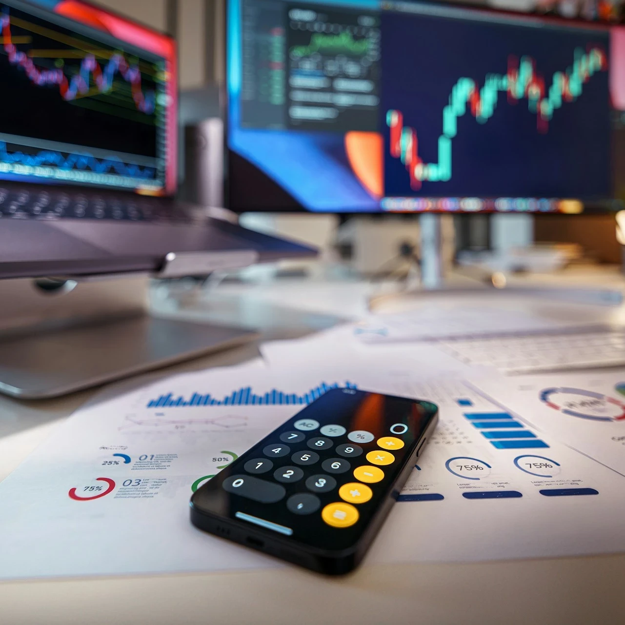 A desk with a calculator and graphs in front of a laptop and a desktop screen.