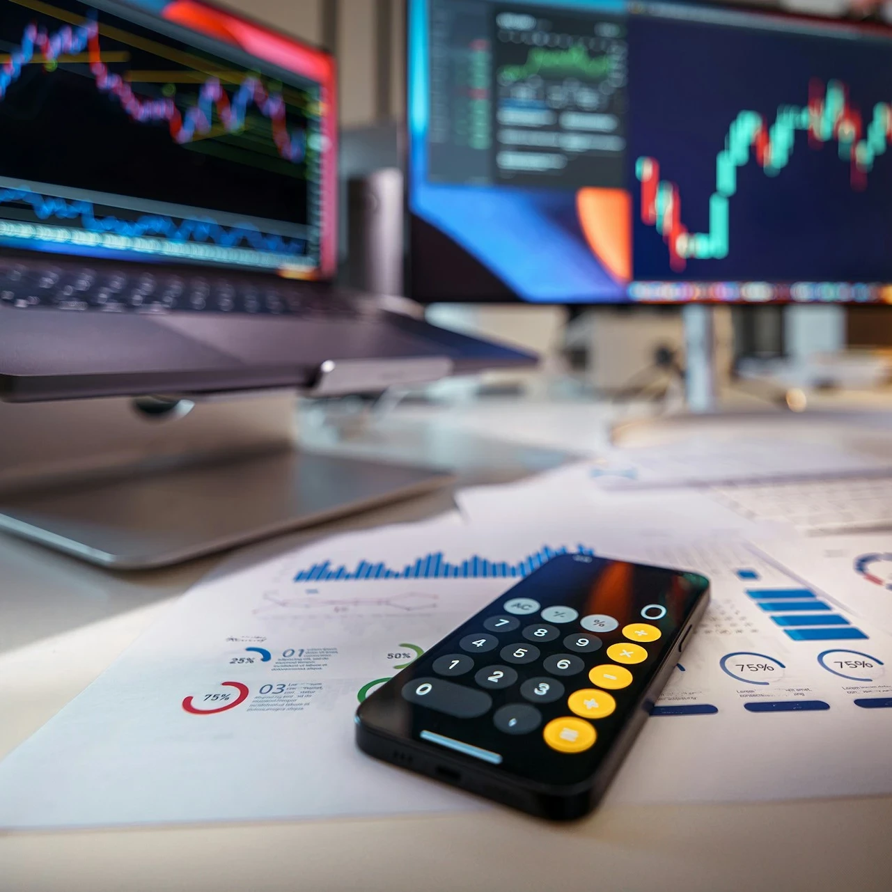 Desk with a laptop and desktop screen on it as well as a calculator app on a smartphone with graphs on paper surrounded it.