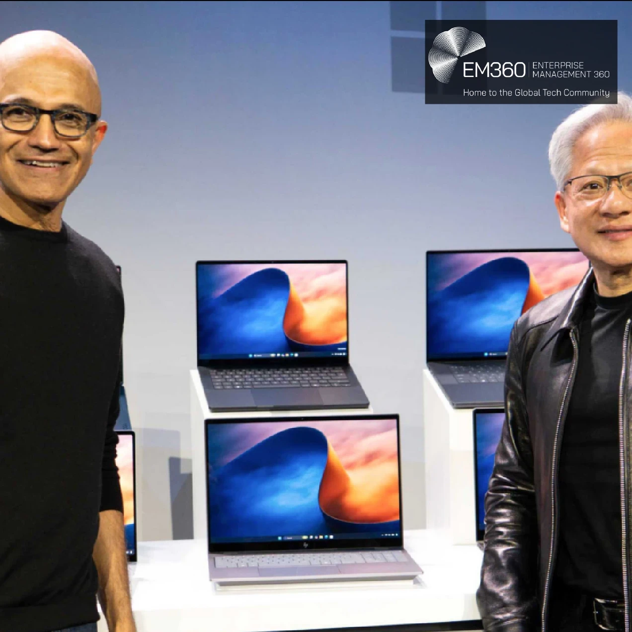 Microsoft and Nvidia executives Satya Nadella (left) and Jensen Huang (right) stand beside several open laptops displaying blue and orange screens, against a grey-blue backdrop with the Microsoft logo. A semi-transparent black overlay across the lower half features the white EM360TECH logo and headline: “Microsoft Gives AI Agents Their Own Security Boundaries in Windows 11.”