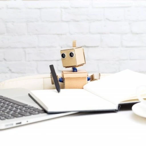 Toy robot pretending to write in a notebook while looking at a laptop