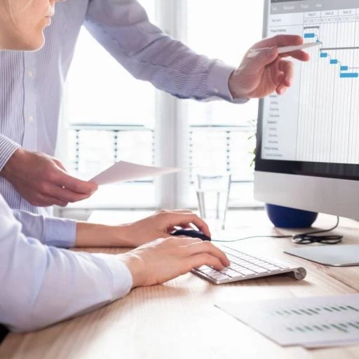 Two businesspeople standing and sitting in front of a desktop computer viewing analytics on a screen.