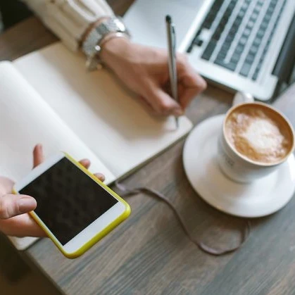 person working on a smartphone, making notes in a book with a coffee and laptop beside them