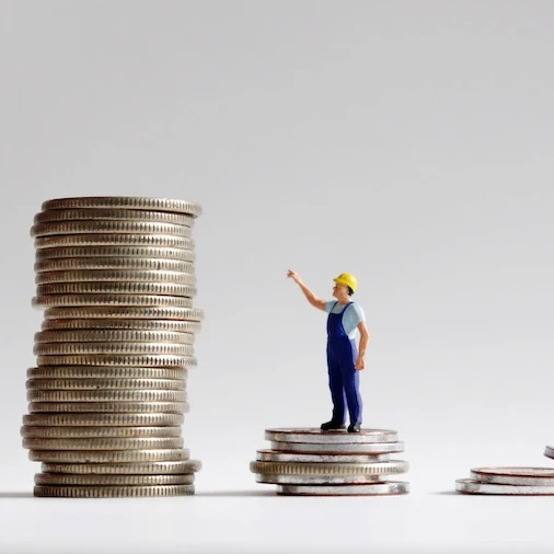 a toy worker in a hardhat standing on a small pile of coins pointing to a larger pile of coins