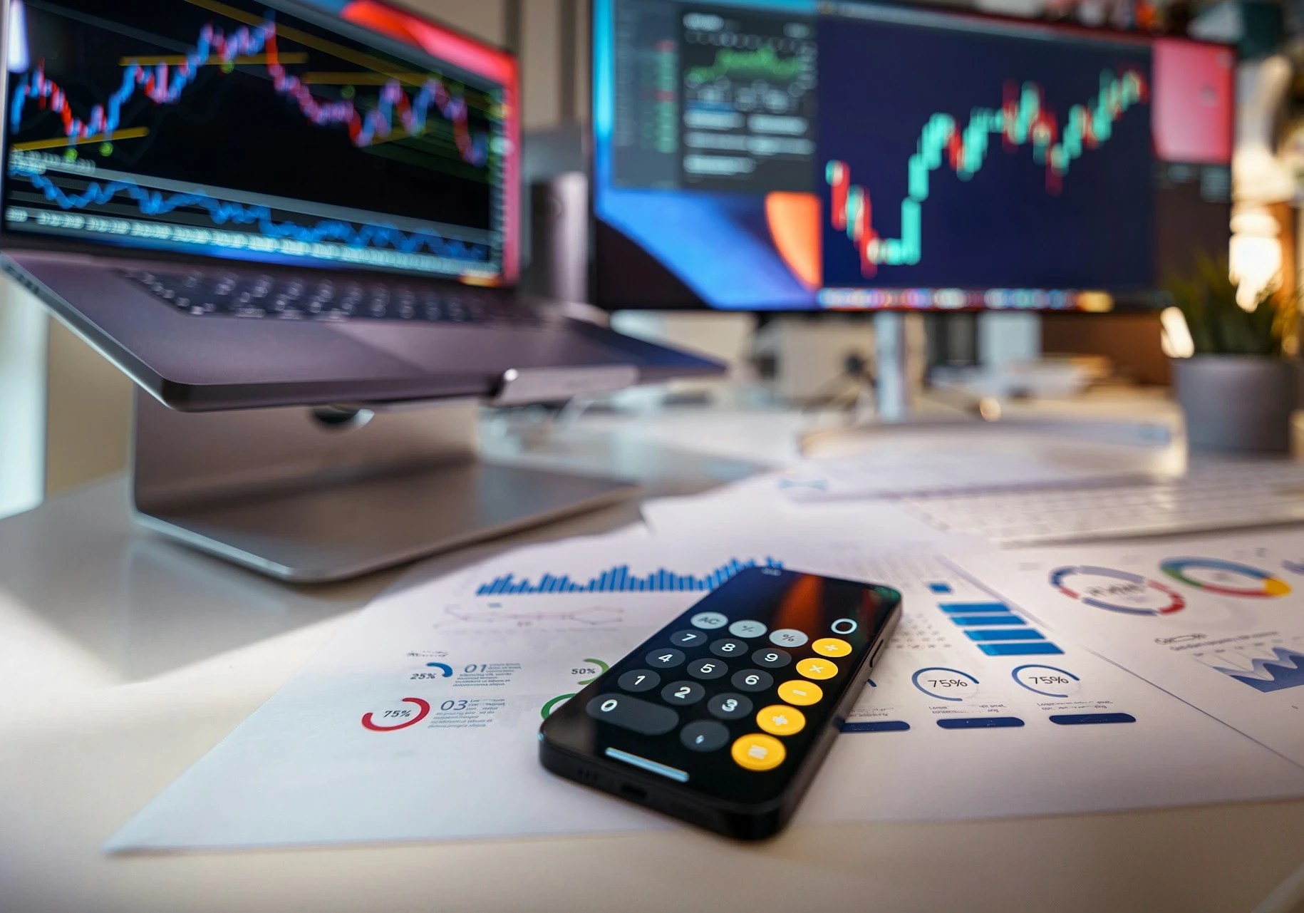 Desk with a laptop and desktop screen on it as well as a calculator app on a smartphone with graphs on paper surrounded it.